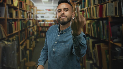 Man shows middle finger toward camera in a library building aisle lined with books and shelves;...