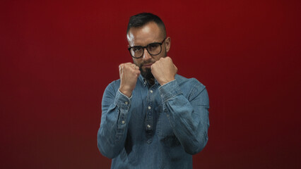 Man young with fists raised punching toward camera wearing denim shirt glasses and beard in studio; determination red grit focus.