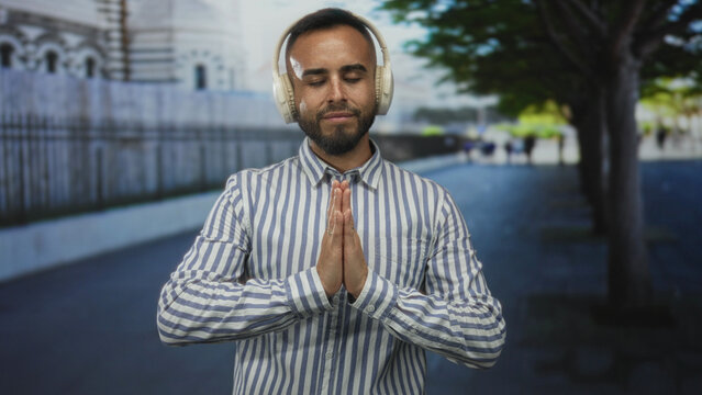 Man wearing headphones, eyes closed, hands pressed in prayer gesture on a city street near trees and stone wall; mindfulness calm.