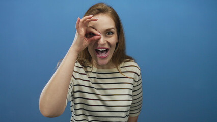 Woman wearing striped shirt frames one eye with binocular hand gesture in blue studio; curiosity...
