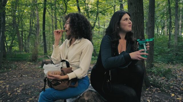A caucasian brunette woman eats a delicious cookie during a snack break.Two female friends are drinking water and tea from a thermos outdoors.