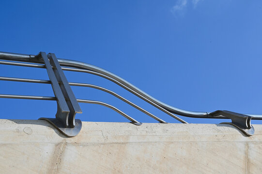 shiny curved stainless steel tubular guard and handrail bolted to chamfered concrete balustrade low stair wall. closeup detail of double L bracket post. horizontal spacer tubes. blue sky background