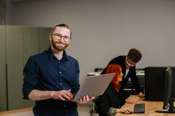 Smiling man holding laptop in modern office environment