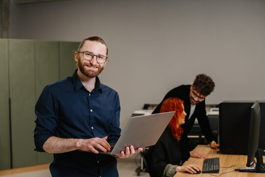 Smiling man holding laptop in modern office environment - Powered by Adobe