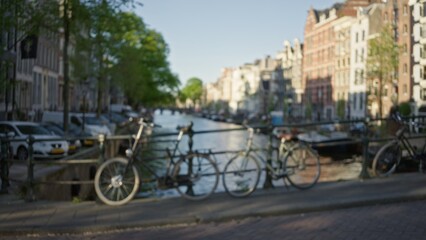 Obraz premium Blurred view of bicycles along the tranquil canals of amsterdam with out-of-focus historic buildings and lush greenery under a vivid blue sky.