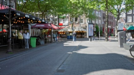 Blurred street scene in a european city with people walking by cafes under a canopy of green trees, creating a vibrant and bustling atmosphere in broad daylight.