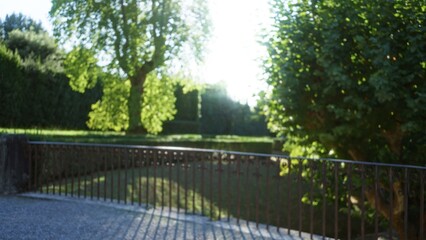 Blurred scene with soft bokeh and defocused iron railing under sunlit trees in a garden, shallow...
