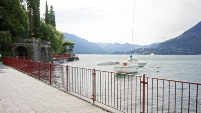Blurred lakeside promenade with red railing, moored sailboat and stone archway in soft bokeh outdoor; background backdrop copyspace.