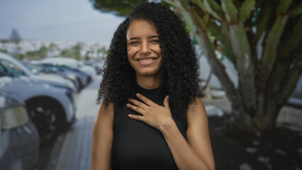 Young hispanic latina woman points index finger while pressing hand on chest in urban parking lot;...