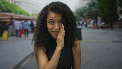Woman with curly hair gesturing secrecy in a bustling city market with blurred background of people shopping outdoors.