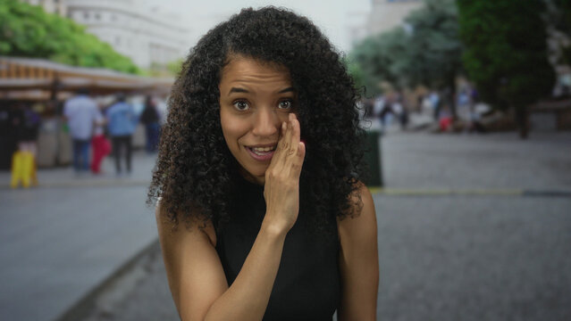 Woman with curly hair gesturing secrecy in a bustling city market with blurred background of people shopping outdoors.