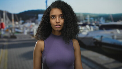 Woman with surprised expression at seaside port with boats and hills in background under clear sky.