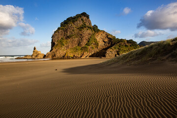 Rugged coastal rock and wind-shaped sand patterns at Piha Beach, New Zealand, under a bright sky.