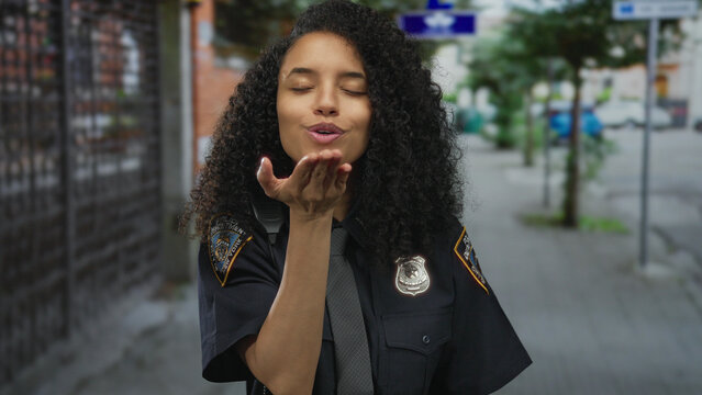 Hispanic policewoman blowing kiss outdoors in uniform on city street with vibrant urban background showcasing confidence and professionalism