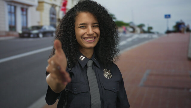 Young hispanic policewoman in uniform extends hand to camera on busy downtown red brick sidewalk by street; trust welcome connection approach. - Powered by Adobe