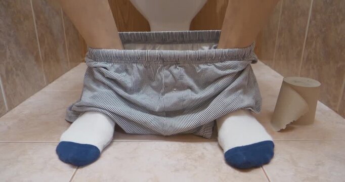 Man sits on toilet sink in restroom and impatiently twitches his legs. Man pushing, can not poop or suffer from constipation. Feet of man sitting on toilet in white socks and with his underwear down