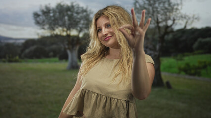 Young blonde woman smiling, giving thumbs up and making ok gesture in green park field with olive tree; confidence.