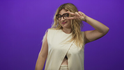 Woman with blonde hair wearing glasses smiling and making a peace sign in a purple studio with sleeveless top and midriff cutout; playful.