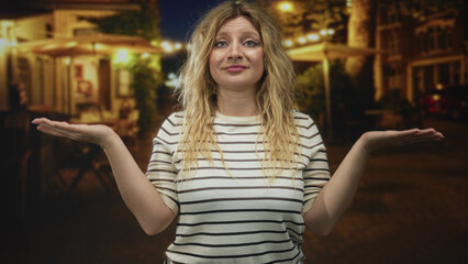 Young blonde woman shrugging with palms up on a dimly lit cobblestone street at night; uncertainty...