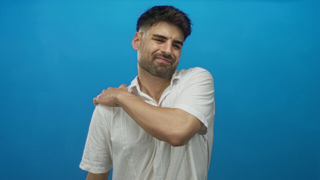 Hispanic young man with hand on shoulder in studio wearing white linen shirt and trimmed beard; discomfort.