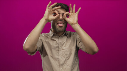 Young hispanic man holds hands to eyes in binocular gesture in pink studio; curiosity exploration wonder.