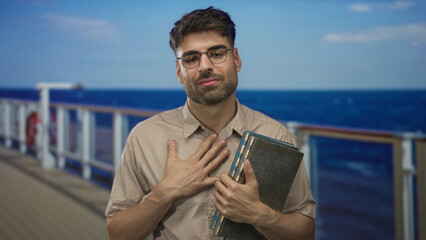 Man wearing glasses holds antique books with hand on chest on wooden boat deck under sunlight;...