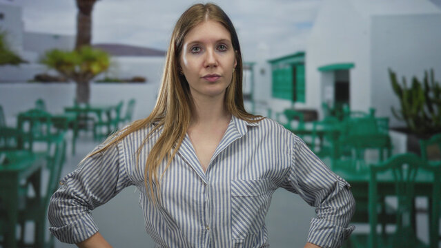 Woman in striped blouse gesturing hand on outdoor restaurant terrace with green chairs and white building facade; confidence.