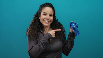 Hispanic woman with curly hair holding a blue ribbon in front of an isolated blue background, smiling confidently suggesting achievement and success.