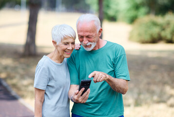 Happy active senior couple having fun using smartphone, taking selfie, or having a video call and wearing sportswear, after having an exercise sport activity outdoors