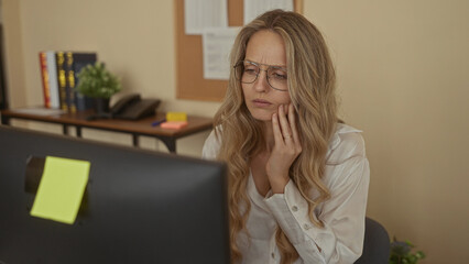 Woman holds cheek in clutch posture in an office as a young blonde at a workplace uses a computer on a desk in an indoor environment.