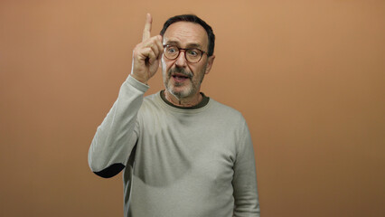 Man posing thoughtfully against brown background in casual outfit and glasses appearing inquisitive and engaged.
