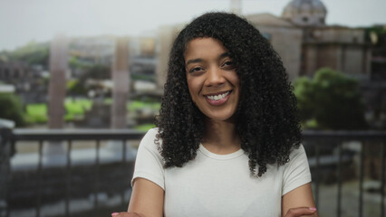 Woman smiling with crossed arms at roman building with stone ruins in bright midday sunlight;...