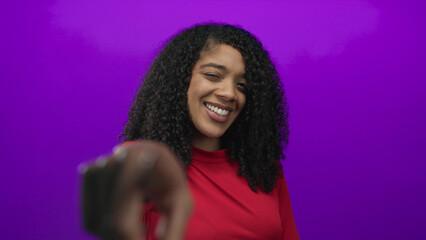 Smiling young african american woman wearing red shirt points finger to camera in studio;...