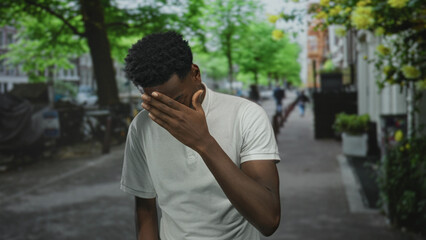 Man covers face with hand on city street under green trees beside blurred buildings and potted...