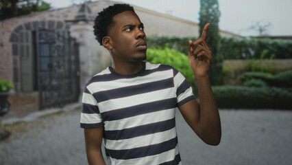 Fototapeta premium Young african american man in navy and white striped shirt points finger at ornate metal gate of stone building entrance under natural light; curiosity.