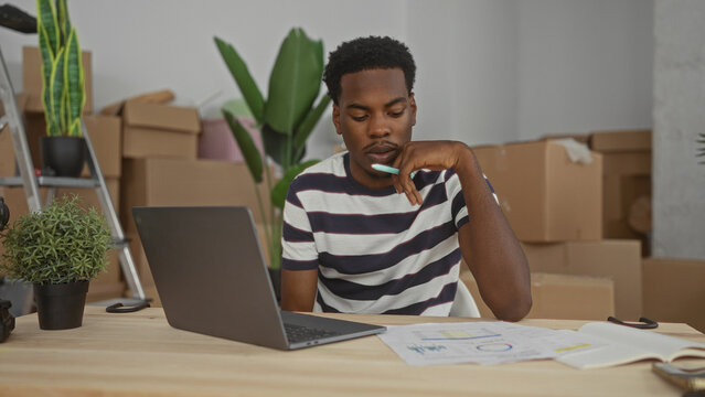 Young african american man typing on laptop in a sparsely furnished building with stacked cardboard moving boxes and potted plants; focus productivity concentration determination.