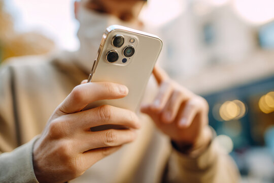 Man wearing face mask using smartphone outdoors focusing on hand holding mobile device with blurred urban background and warm sunlight reflections
