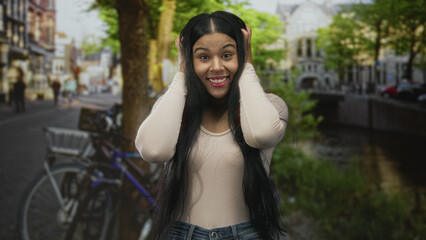 Woman covering her ears with hands by a canal in a sunny city street lined with bicycles, smiling...
