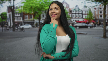 Woman in green cardigan with hand on chin and slight smile looking upward on busy cobblestone street; contemplation.
