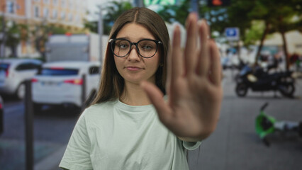 Teenage girl wearing glasses shows stop hand gesture on bustling city street with parked cars; boundary refusal assertion defiance.