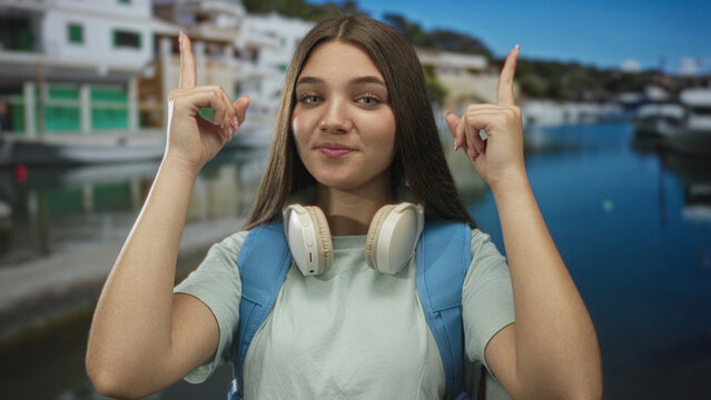 Girl with backpack and headphones points finger upward at moored boats in port under bright sunlight; curiosity.