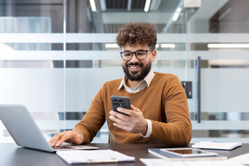 Young man working at a desk in a modern office, smiling while checking his smartphone and simultaneously typing on a laptop, representing multitasking and digital communication