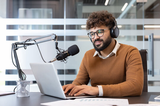 Young man wearing headphones and glasses sitting at a desk, typing on a laptop and speaking into a professional microphone, creating audio content for a podcast or online stream