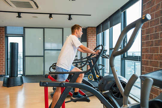 Man working out on an elliptical trainer in modern gym with city view. Male doing cardio training on stationary exercise bike machine in fitness studio. Fitness, wellness, healthy lifestyle