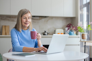 Senior woman smiling, enjoying a cup of coffee while typing on her laptop at the kitchen table, embodying remote work and technology for aging individuals
