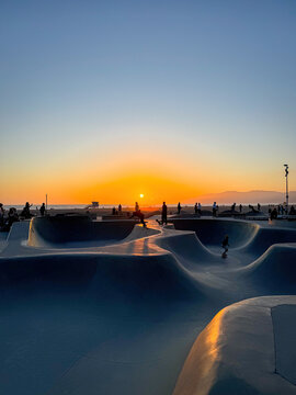 Skateboarders ride the Venice Beach Skatepark in Los Angeles, California, as the sun sets over the Pacific Ocean, the silhouettes and golden reflections create a dramatic and atmospheric coastal scene