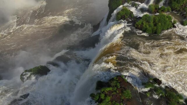 Rushing water crashes down Iguazu Falls sending up clouds of mist in Argentina. Aerial view o highlights immense force as river swirls below. Lush green patches dot the dramatic cliffside scenery