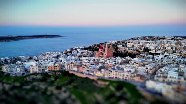 Tilt-shift Drone orbit around Mellieħa Parish Church &mdash; miniature diorama effect with warm sunset light over the coastal town and Mediterranean sea