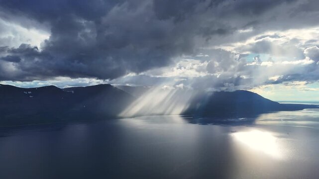 Rain core and slant light meet over the mountain wall by Lake Lama, forming bright veil on the water. Mist curls along the ridge, highlighting the raw Siberia landscape and timeless volcanic plateau
