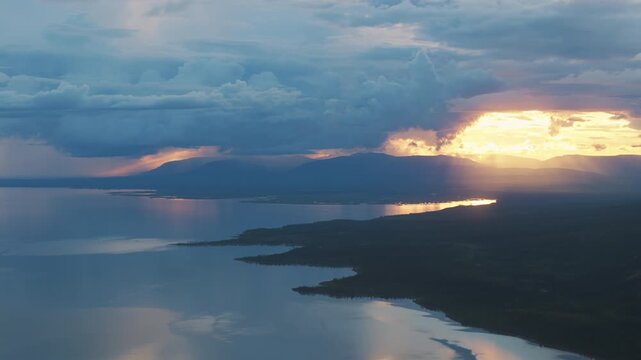 Sun breaks through storm cloud and lights the bays of Lake Lama, painting reflections on distant ranges. Dramatic weather and arctic clarity create a compelling travel scene on the Putorana Plateau